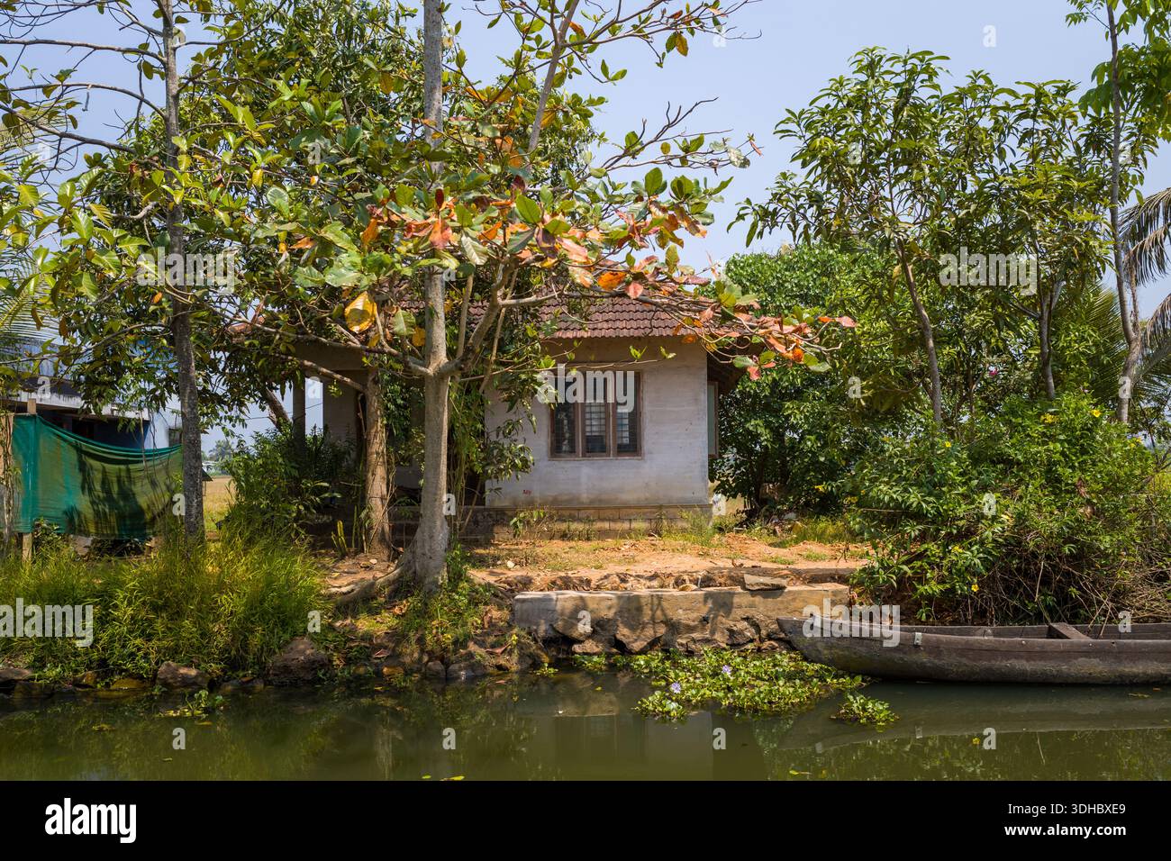 Alappuzha, India - 9 marzo 2023: Una piccola casa con un tetto piastrellato è situata sulla riva delle backwaters, circondata da alberi e vegetazione, con una canoa in legno ormeggiata sul bordo dell'acqua. Foto Stock