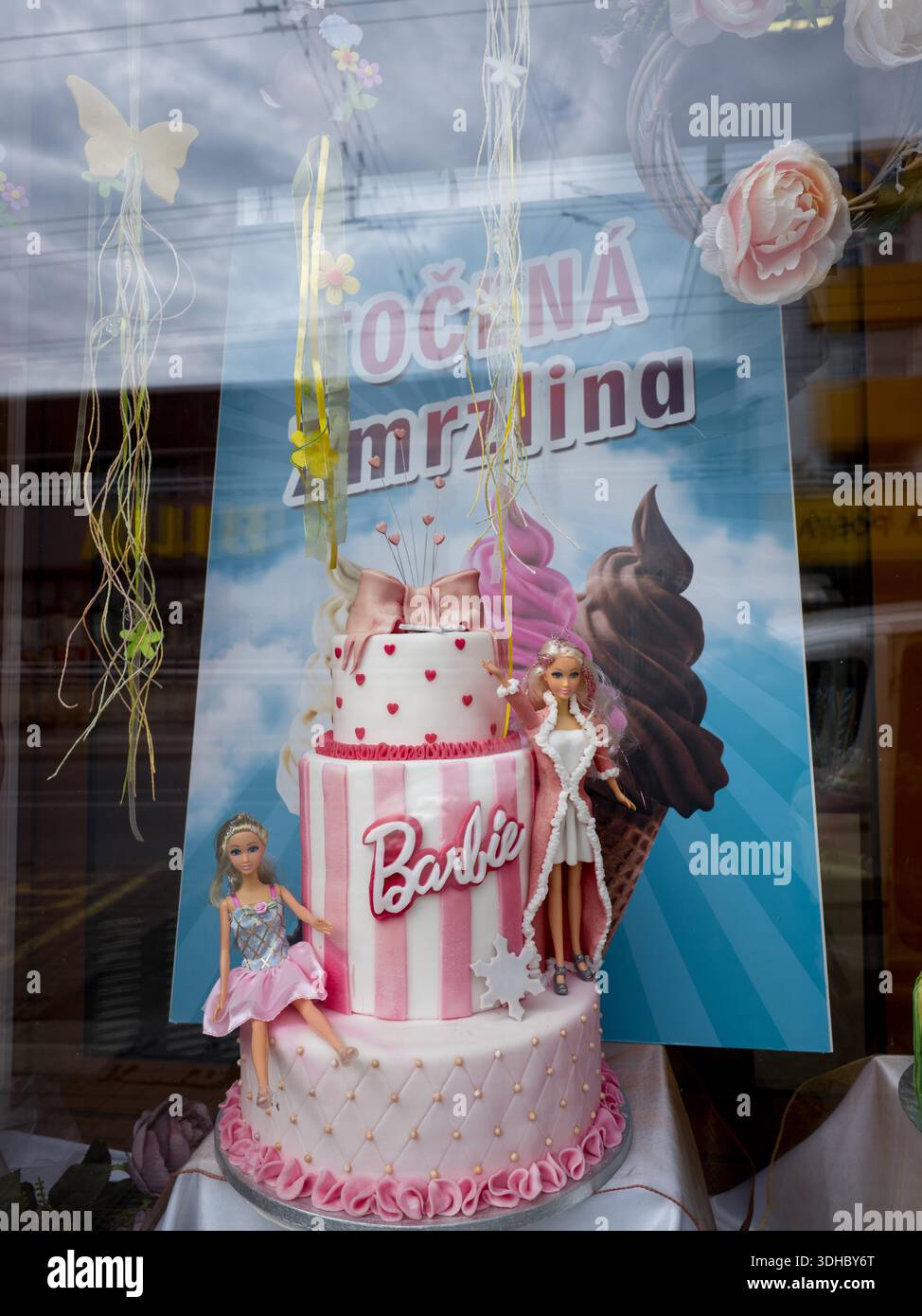 Una torta a tema Barbie rosa e bianca con bambole esposte in una vetrina vicino a una pubblicità di gelati cechi. Foto Stock