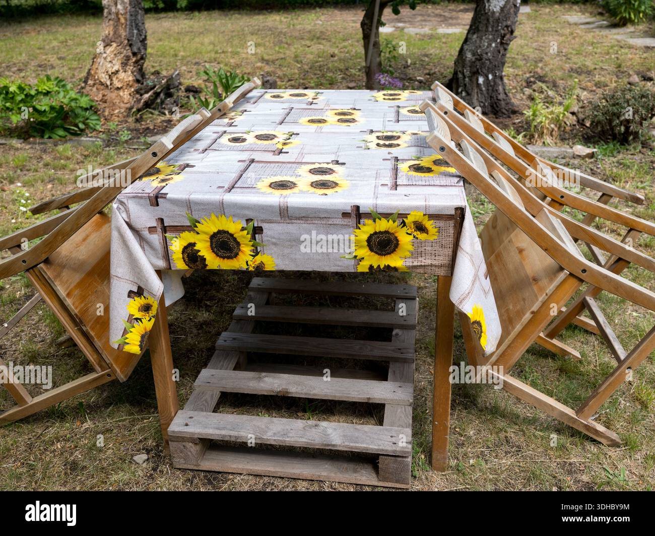Un tavolo da giardino in legno ricoperto da una tovaglia con motivo girasole, con quattro sedie inclinate contro i lati all'esterno. Foto Stock