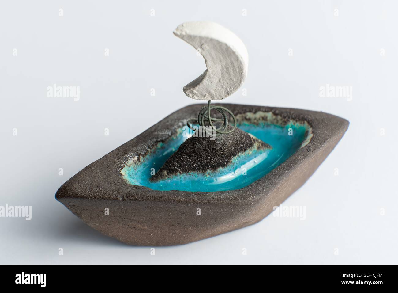 Questo pezzo di artigianato presenta una piccola barca realizzata in materiale scuro. Ha una luna crescente leggera in cima e acqua blu intorno a una forma centrale. Foto Stock