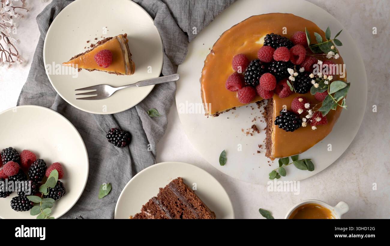 Una deliziosa torta con strato di cioccolato condita con una liscia glassa di caramello e guarnita con lamponi e more. La scena è caratterizzata da un elegante design piatto Foto Stock