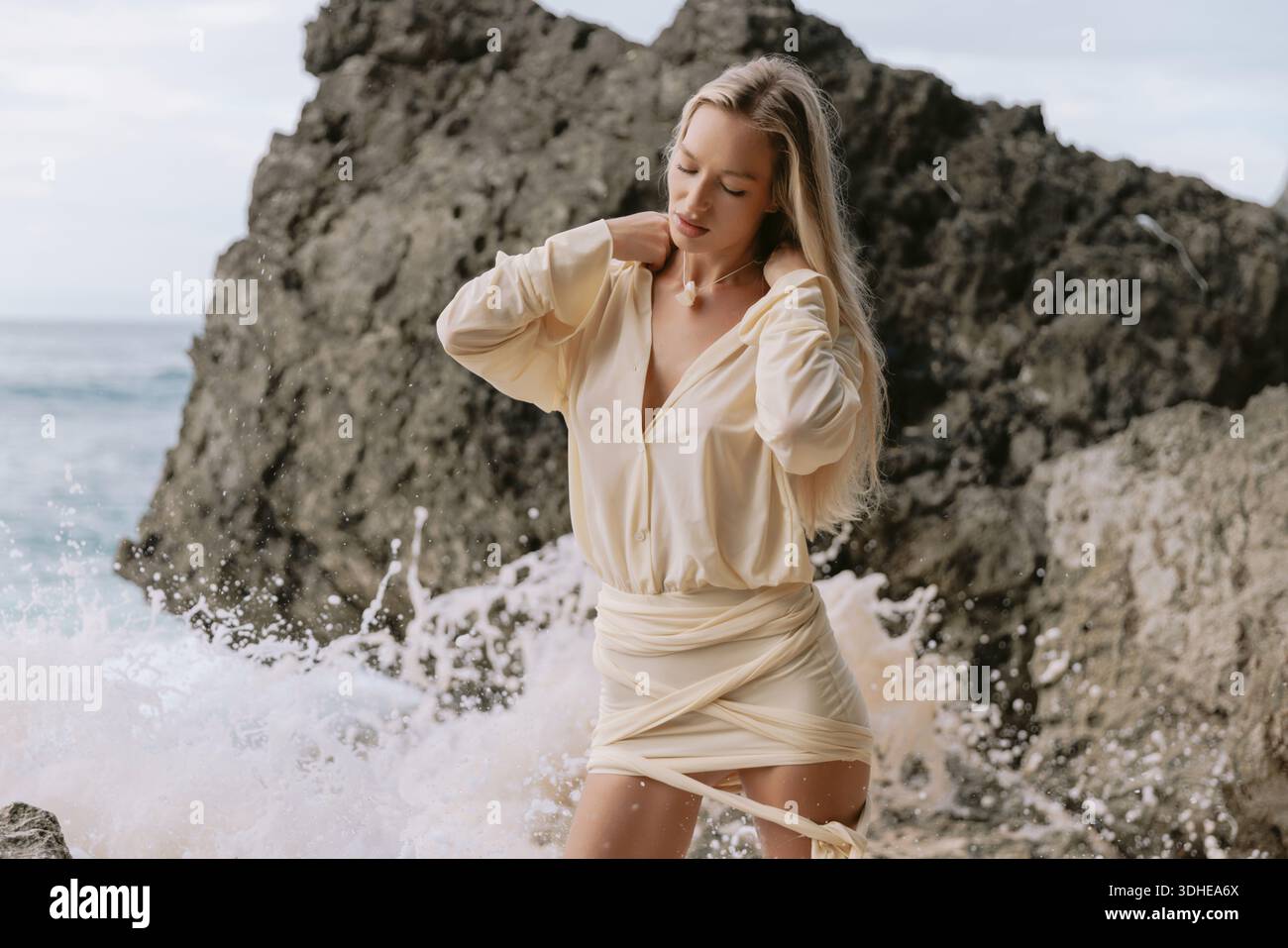 Elegante donna bionda in abito beige in piedi su una spiaggia rocciosa con onde dell'oceano che spruzzano Foto Stock