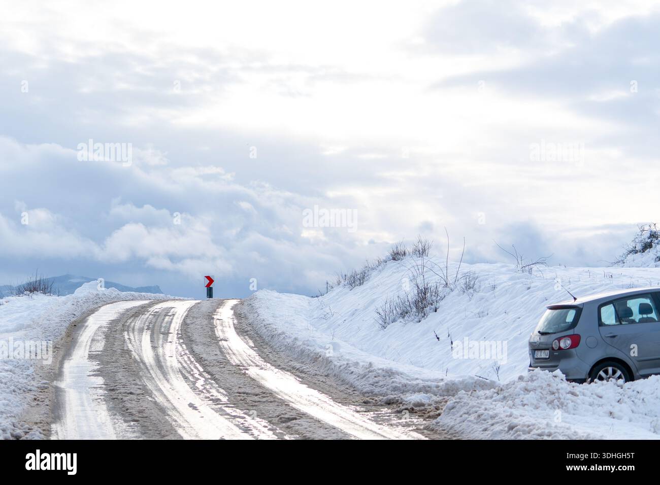 Un veicolo è parcheggiato a lato di una strada innevata e tortuosa. Il paesaggio presenta montagne e un cielo nuvoloso. La neve copre il terreno e le parti della r Foto Stock