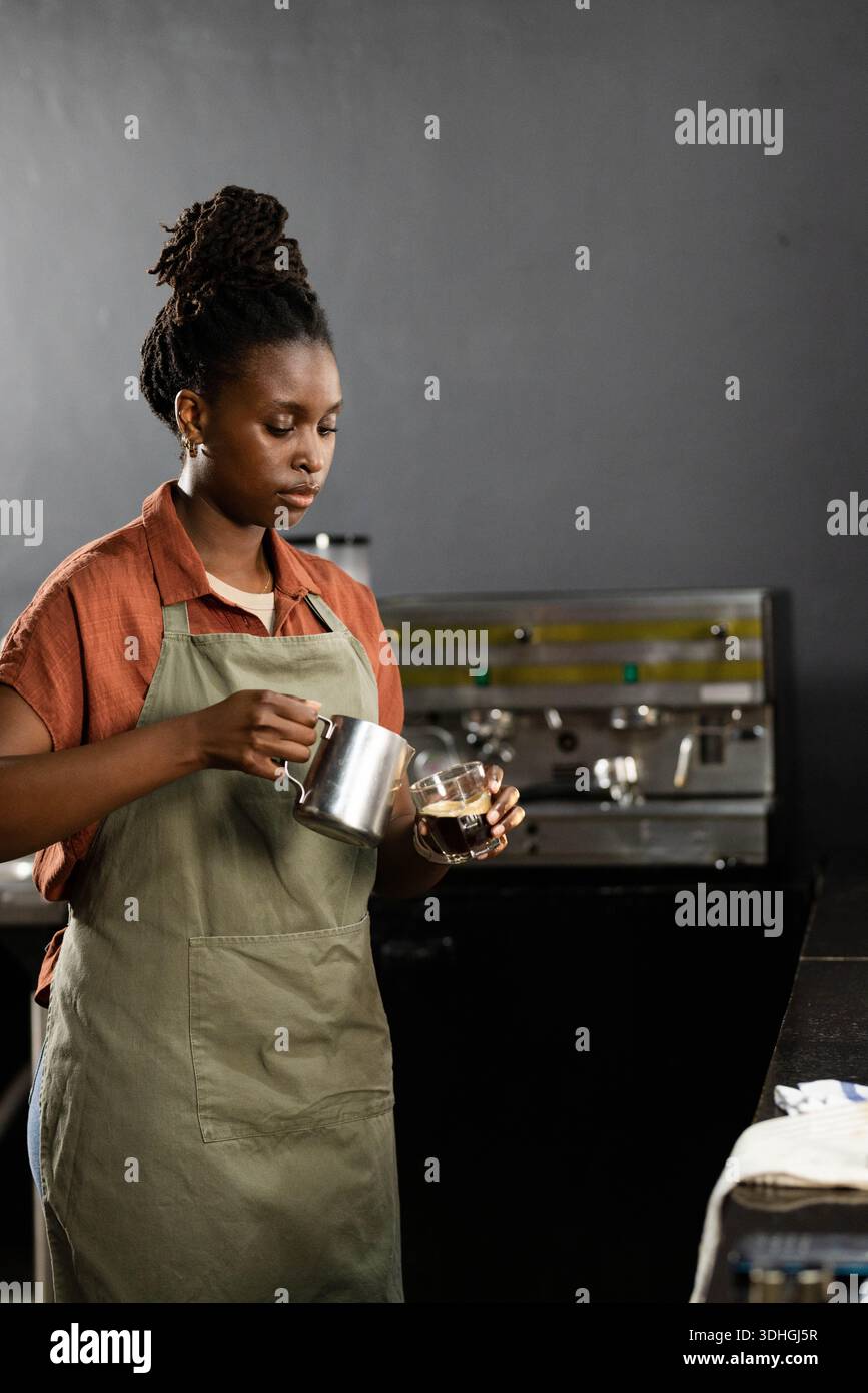 Donna africana che indossa un grembiule verde che versa latte in una tazza di vetro al bar vicino alla macchina per espresso Foto Stock
