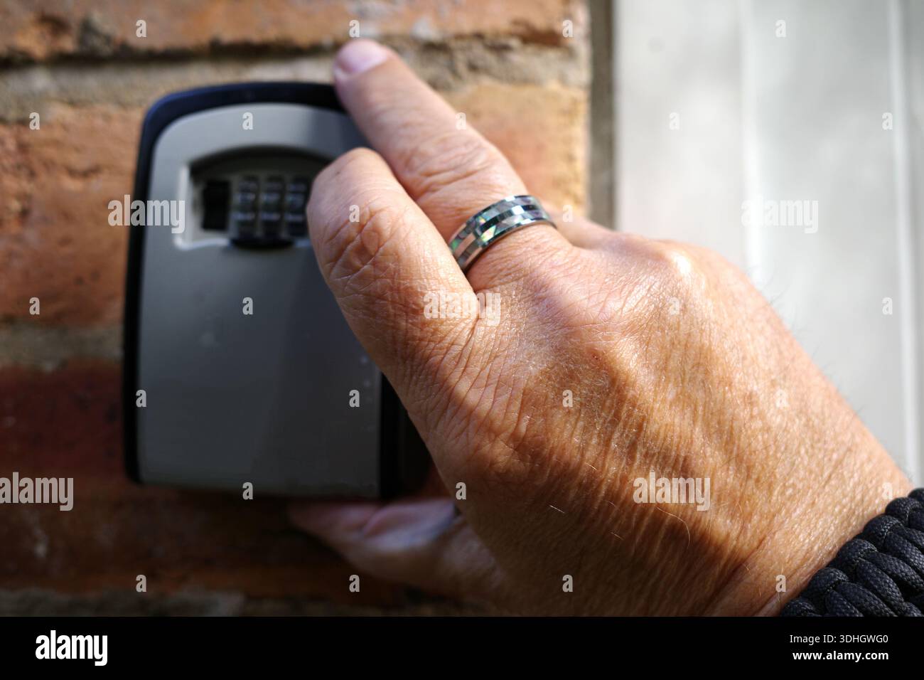 La mano di una persona che indossa un anello si vede aprire una cassetta per chiavi montata su una parete di mattoni. La cassetta portachiavi è grigia con una finitura nera e presenta una combinazione numerica Foto Stock