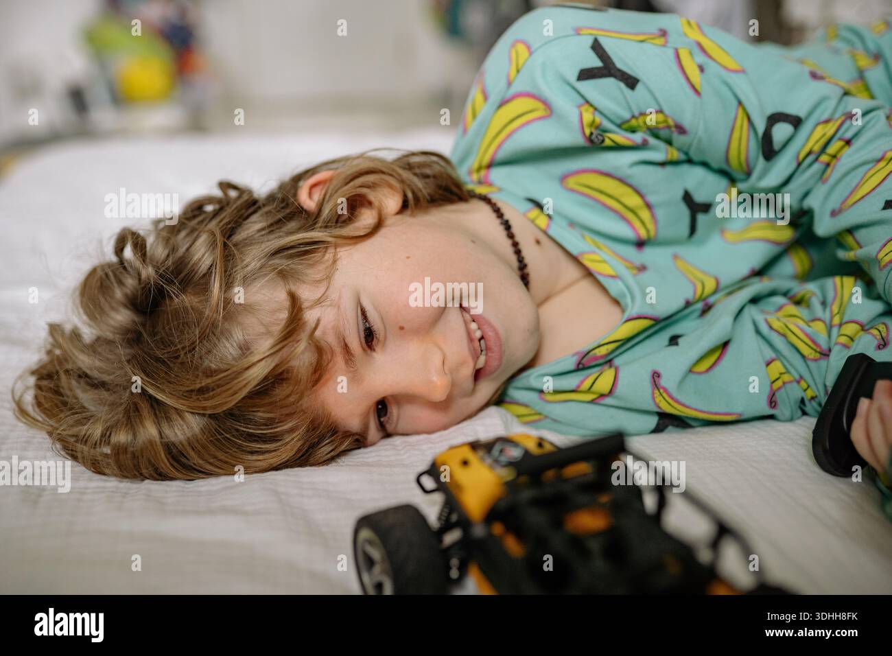 Un ragazzo giace sullo stomaco nella sua camera da letto. Sorride mentre gioca con un'auto giocattolo, indossando pigiama colorati. La camera presenta decorazioni giocose visibili e. Foto Stock