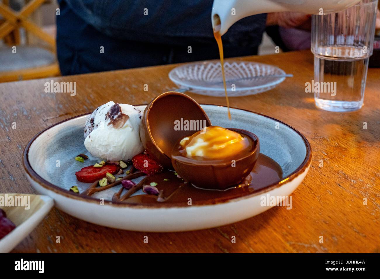 Uovo di cioccolato con gelato e frutta sul piatto, caramello liquido sulla caraffa, bicchiere d'acqua sul tavolo. Preparazione del dessert in tavola. Foto Stock