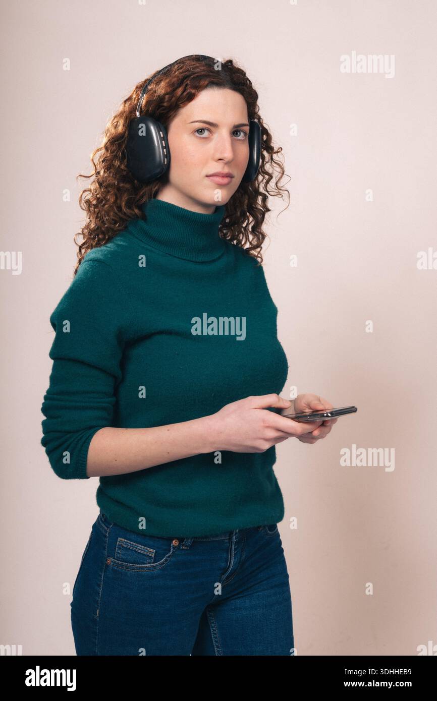 Una giovane donna con capelli rossastri ricci, con cuffie e maglione verde, tiene in mano uno smartphone e ascolta la musica in uno studio. Foto Stock