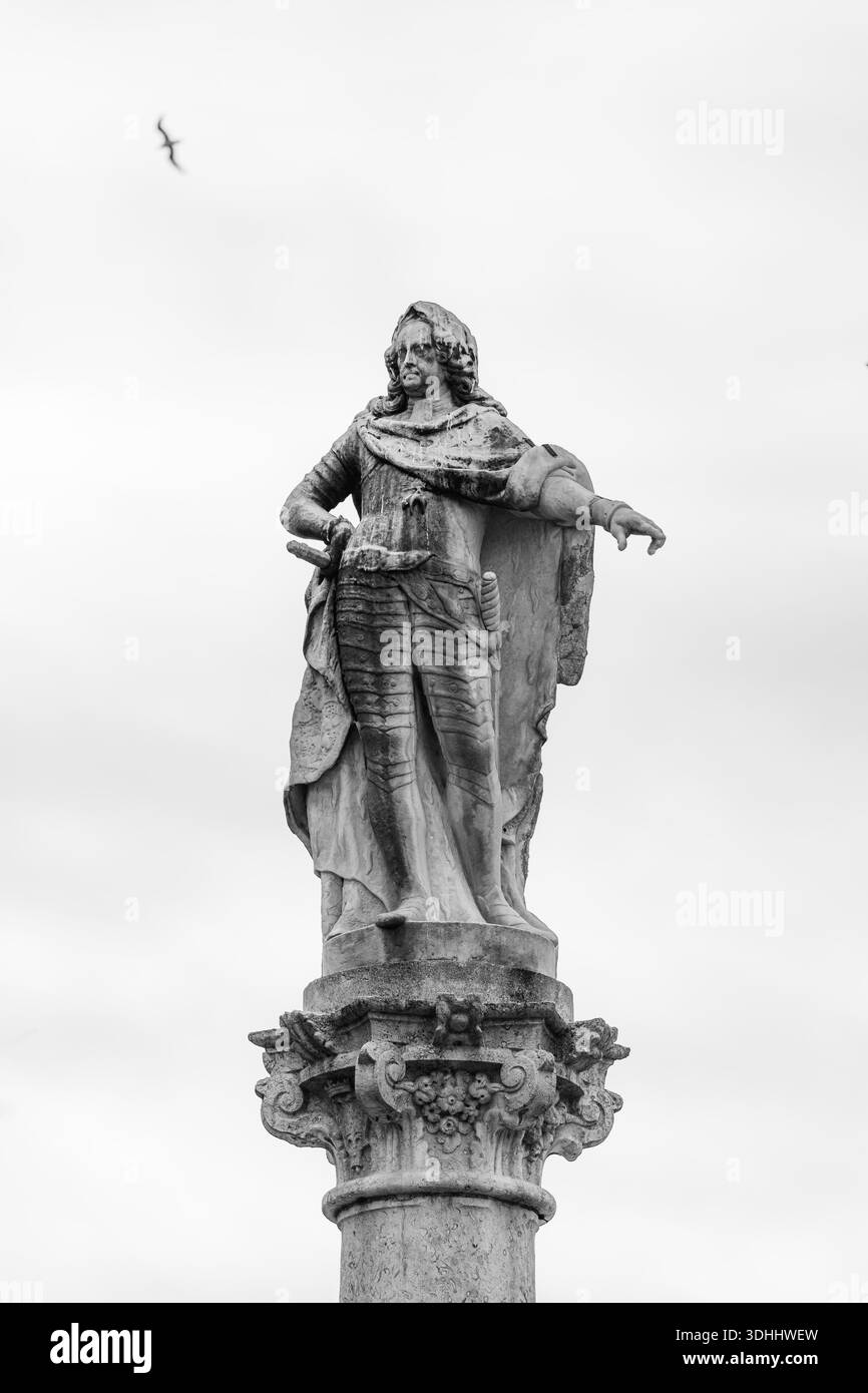 Foto d'arte in bianco e nero di una storica statua in pietra che si erge in cima a una colonna classica ornata, isolata contro un cielo luminoso e minimale. Il co Foto Stock
