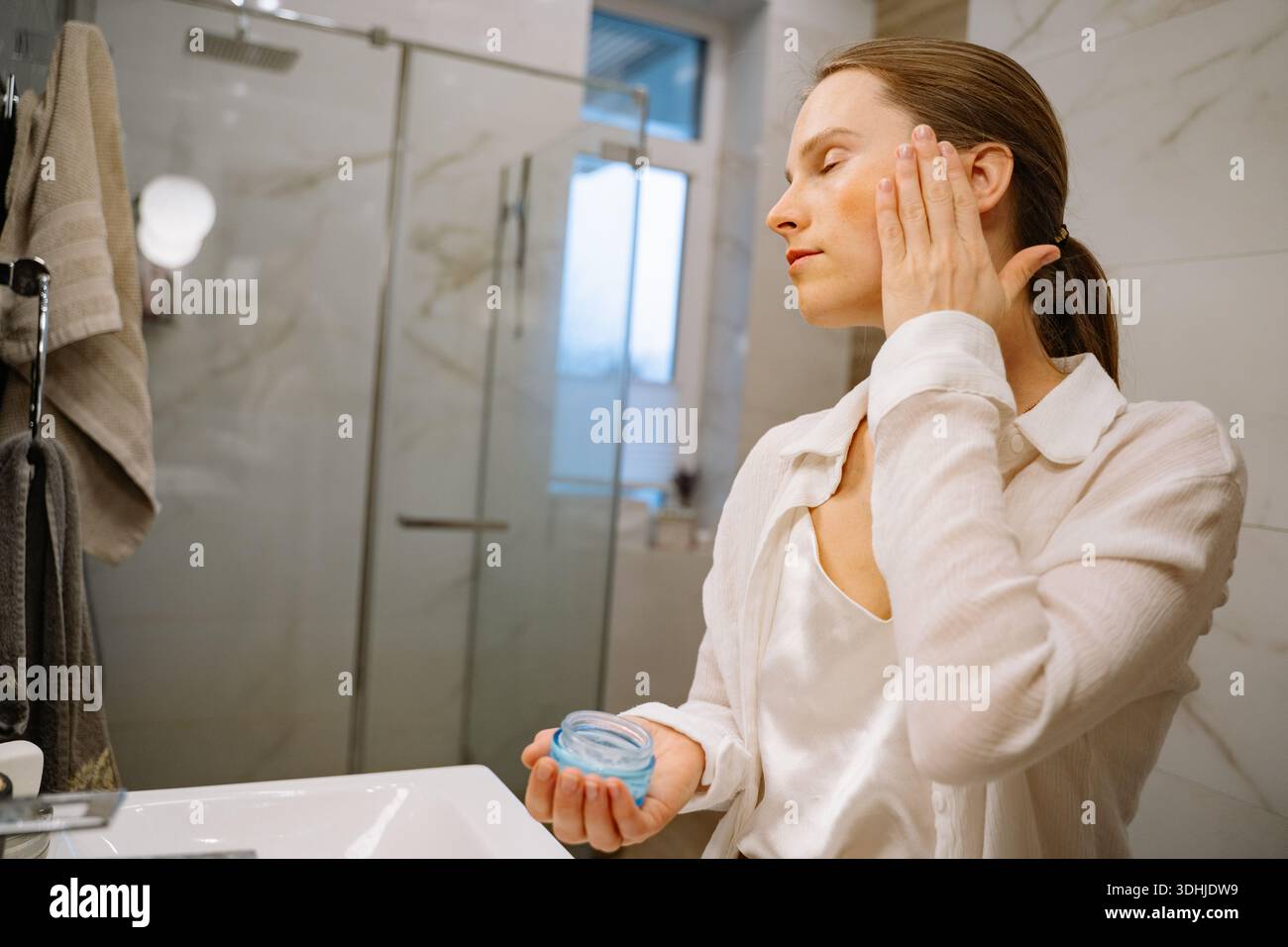 Una donna sta di fronte a un lavandino del bagno, applicando un prodotto per la cura della pelle al viso. Il bagno ha un design moderno con luce naturale Foto Stock