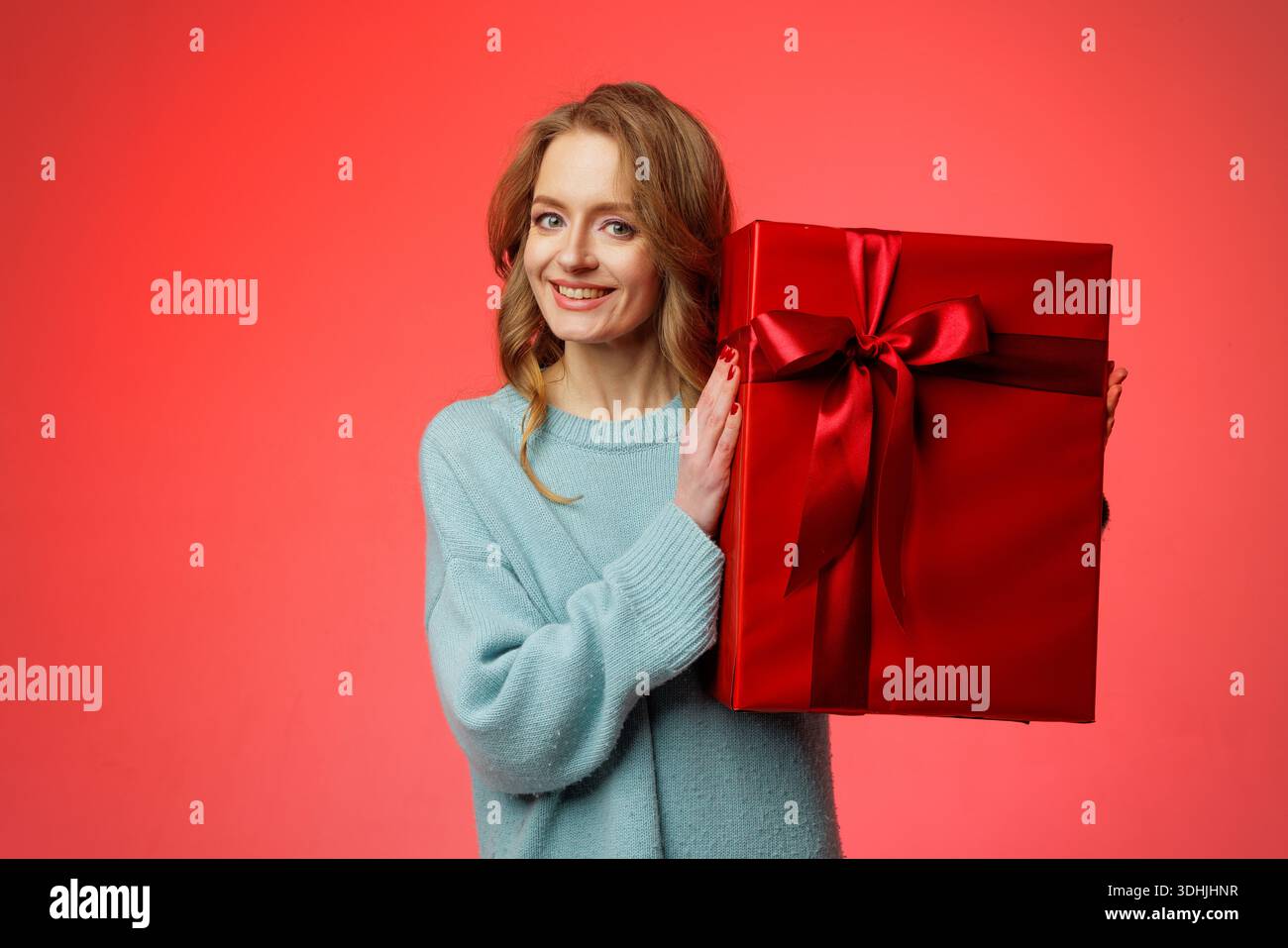 La donna sorridente presenta una grande confezione regalo rossa con nastro su sfondo rosso studio, ideale per banner e-commerce o promozioni di saldi natalizi. Foto Stock