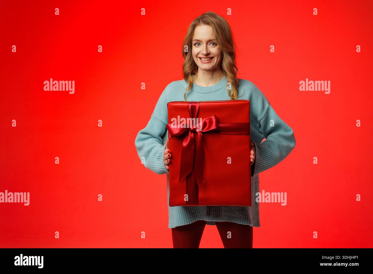 Donna sorridente in maglione con grande confezione regalo confezionata sullo sfondo rosso dello studio, ideale per banner di siti Web o annunci social per promozioni natalizie. Foto Stock