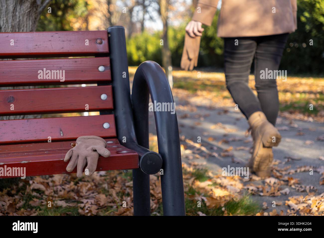 Primo piano di un guanto marrone chiaro su una panchina rossa in legno del parco, con una persona sfocata che si allontana sullo sfondo, incarnando i temi di una passeggiata autunnale Foto Stock