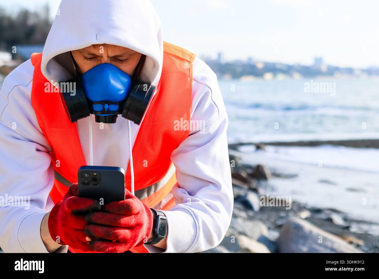 Indossa il respiratore e i guanti utilizzando lo smartphone sulla costa rocciosa con vista sul mare e sugli edifici urbani distanti. Foto Stock