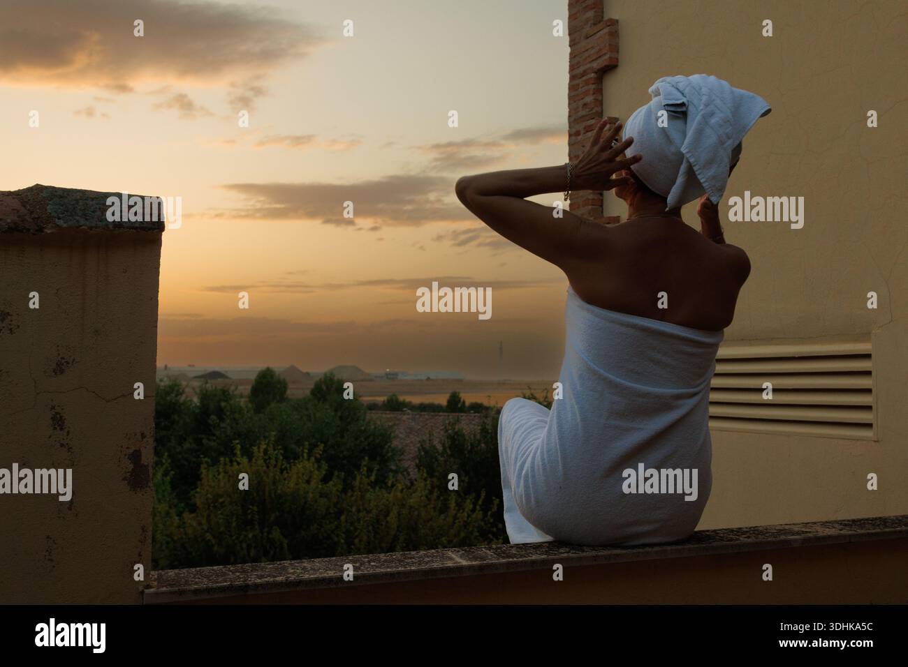 donna seduta a parete con un asciugamano al tramonto, viaggiatore che si adatta mentre guarda verso colline lontane, Foto Stock