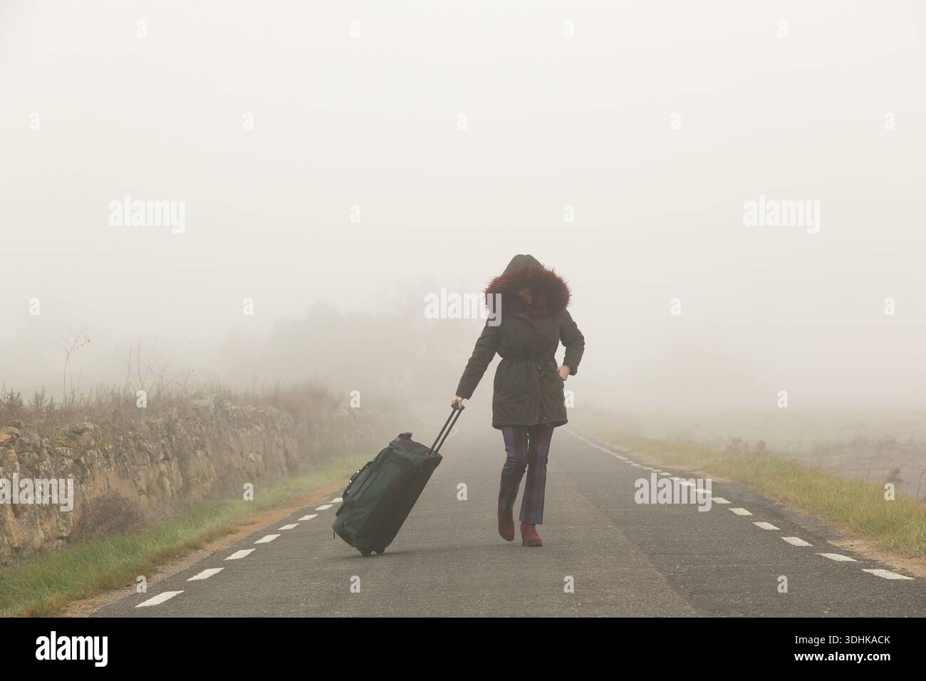 donna bianca che si allontana con la valigia lungo una strada vuota, cappuccio di pelliccia sollevato, andatura costante, trasporto bagagli al traino, Foto Stock