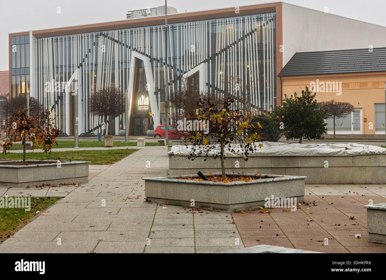VINKOVCI, CROAZIA, 12.13.2025: Una biblioteca cittadina con una facciata in vetro e ingresso scultoreo si erge accanto a una piazza pavimentata con fioriere e foglie autunnali, con Foto Stock