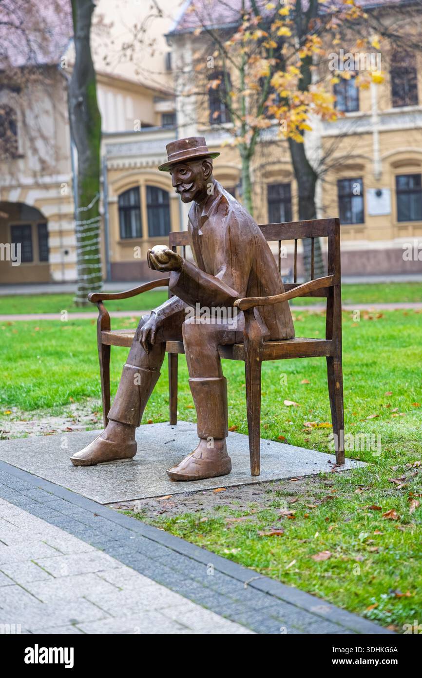 VINKOVCI, CROAZIA, 12.13.2025: Una statua di bronzo di un uomo che indossa un cappello siede su una panchina di legno in un parco cittadino, tenendo in mano una mela. Foglie autunnali, alberi, AN Foto Stock