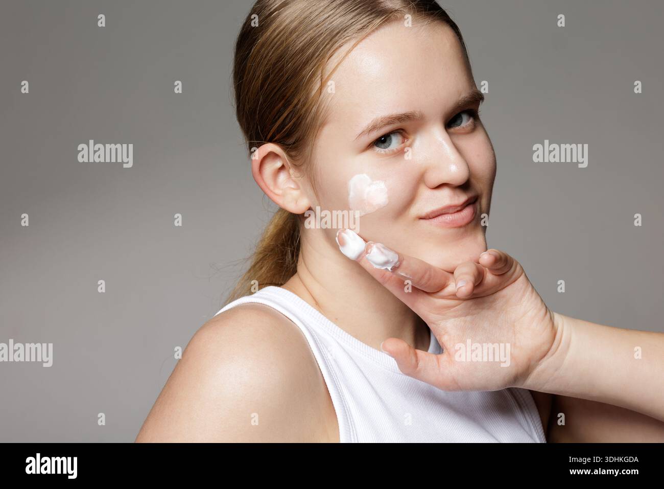Ritratto in studio di una giovane donna che applica crema facciale sulla guancia. Sfondo grigio minimo con spazio di copia per banner, sito Web o blog per la cura della pelle. Foto Stock