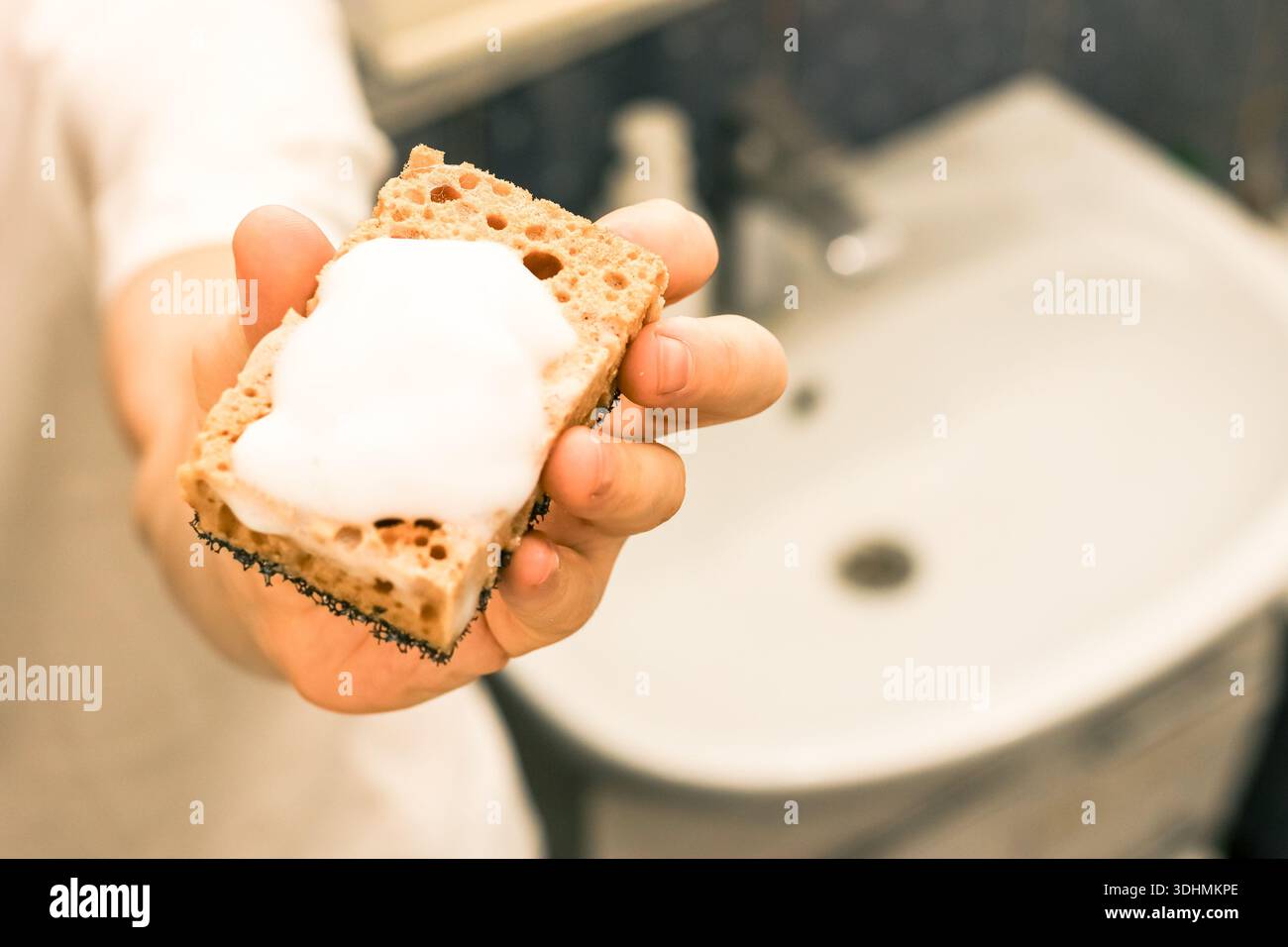 Foto dettagliata di dita che tengono saldamente in mano una spugna gialla con abbondante schiuma bianca, su uno sfondo sottile del bagno. Foto Stock