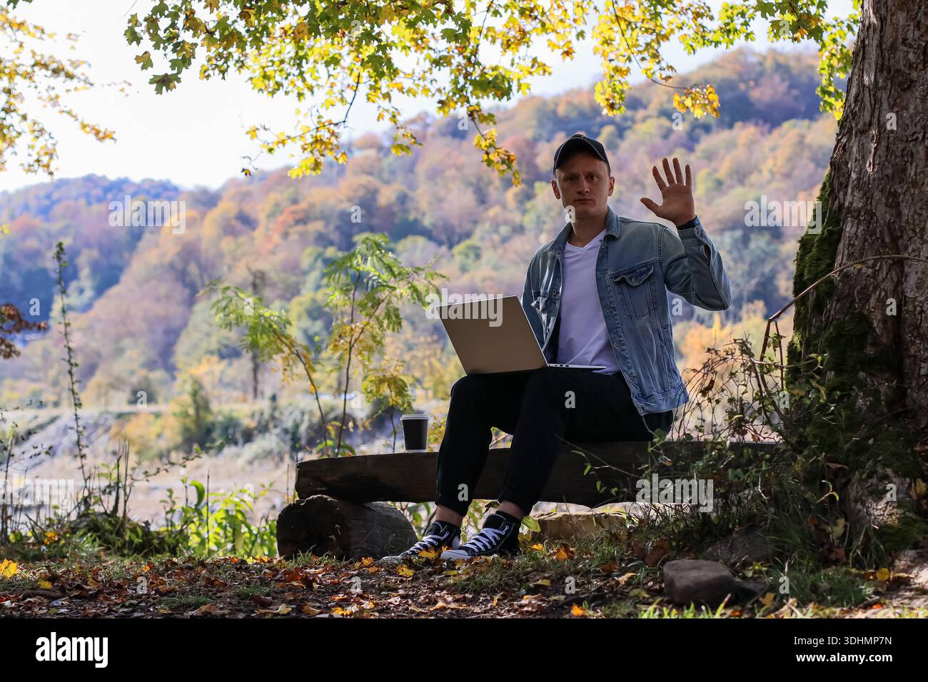 L'uomo cordiale ondeggia la mano, impegnato con il computer portatile e il caffè su una panchina rustica, con i vibranti alberi autunnali e sullo sfondo delle montagne Foto Stock