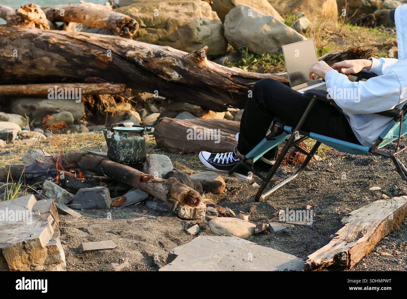 Momento accogliente del campo: Gesti individuali dai capelli chiari espressivi sul laptop in sedia blu, spaccature con vaso, fiume e paillettes in morbida serata Foto Stock