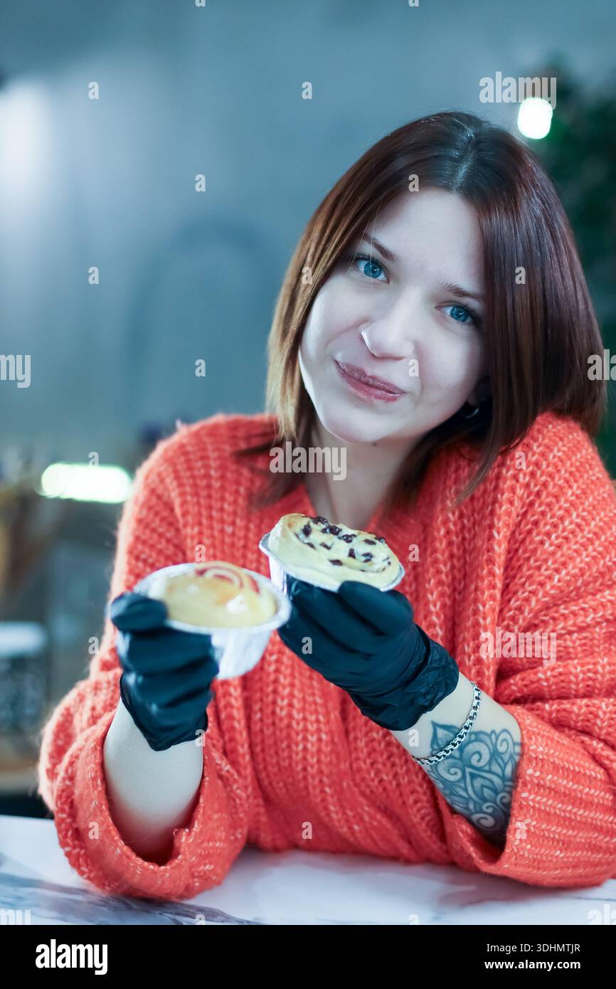 Donna che guarda la macchina fotografica mentre tiene in mano due piccoli articoli da forno con guanti neri, uno con spruzzi di cioccolato Foto Stock