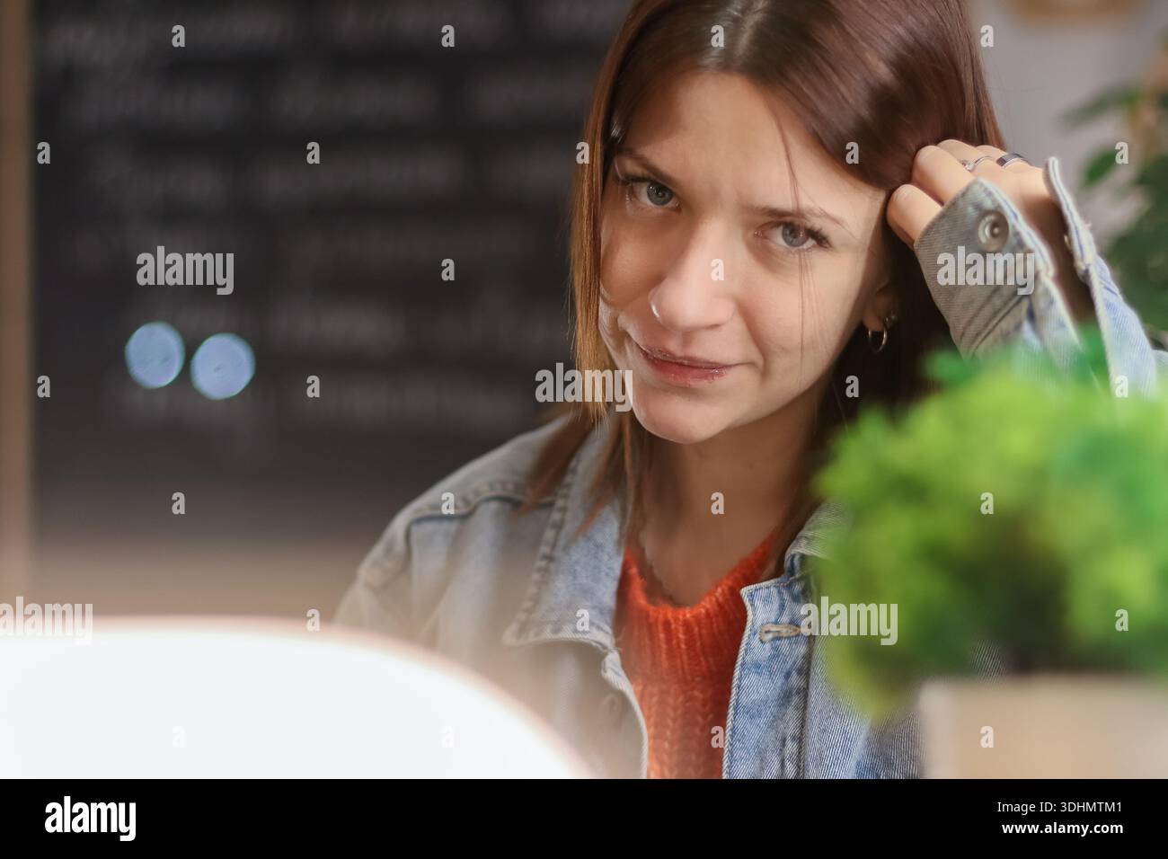 Giovane donna in giacca di denim che guarda con un sorriso sottile mentre legge un libro al tavolo del caffè con le piante Foto Stock