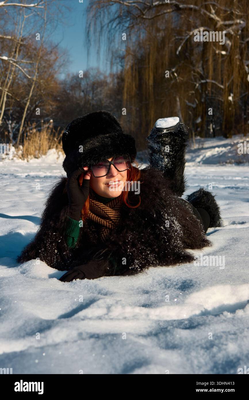 In una scena invernale soleggiata, una donna radiosa con i capelli rossi ardenti si rilassa nella neve incontaminata, il suo viso brilla di gioia. E' avvolta dentro Foto Stock