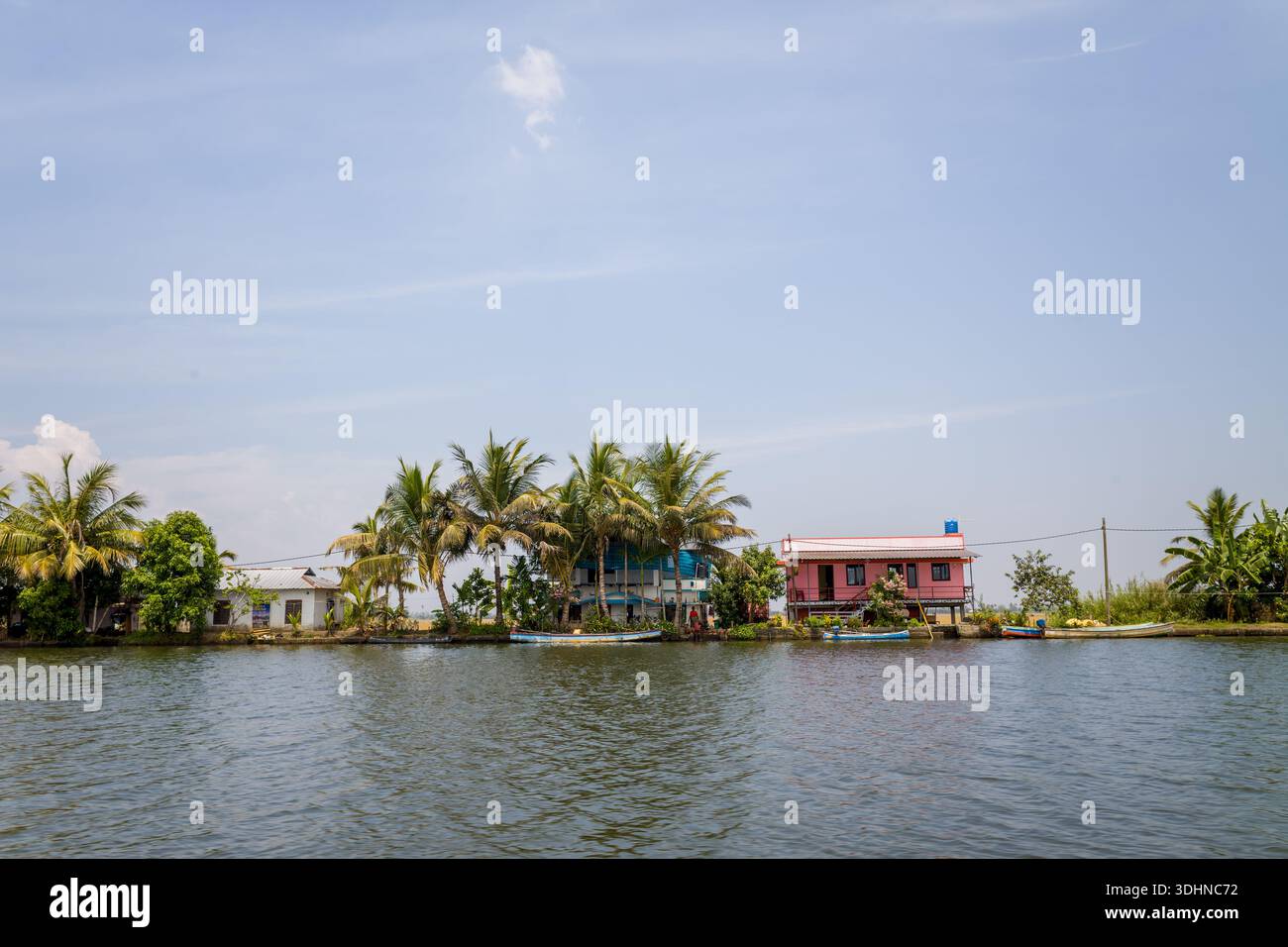 Alappuzha, India - 6 marzo 2023: Diverse case lungo il fiume con esterni colorati e palme circostanti sono visibili lungo le acque secondarie sotto un cielo limpido. Foto Stock