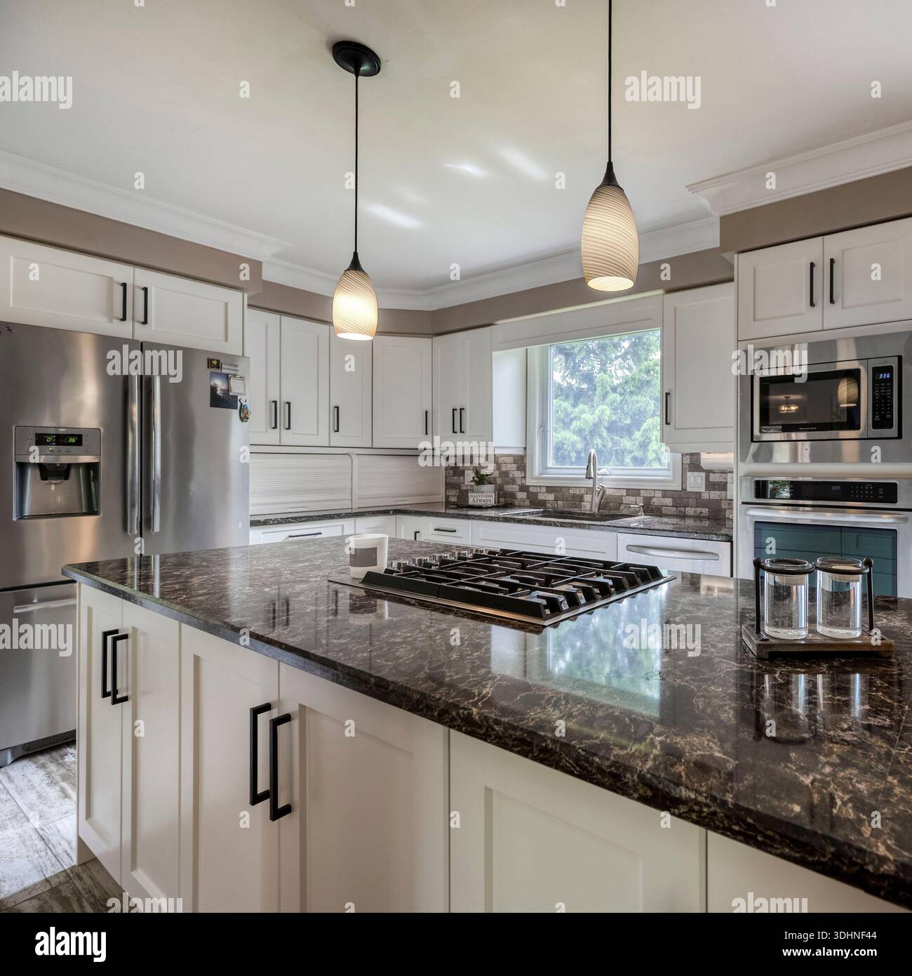 Cucina moderna e luminosa con mobili bianchi, ripiani in pietra, elettrodomestici in acciaio inox, piano cottura a gas, illuminazione a sospensione, e luce naturale. Foto Stock