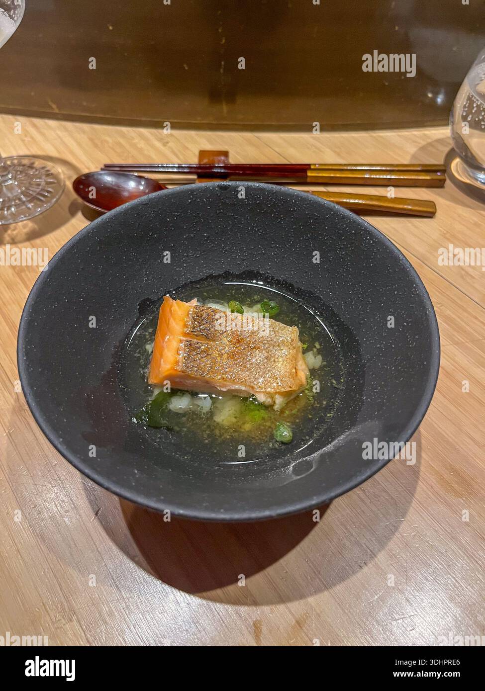 Il filetto di salmone scottato in padella dalla pelle croccante galleggia in un brodo limpido con decorazioni verdi, servito in una ciotola nera su un tavolo di legno chiaro. Foto Stock