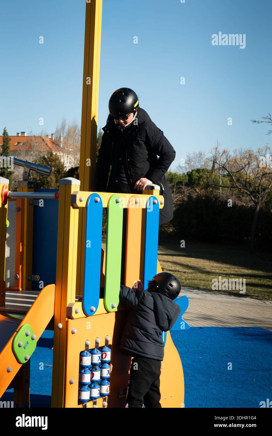 Un bambino che indossa un casco e una giacca invernale arrampicandosi su un parco giochi colorato con un adulto Foto Stock