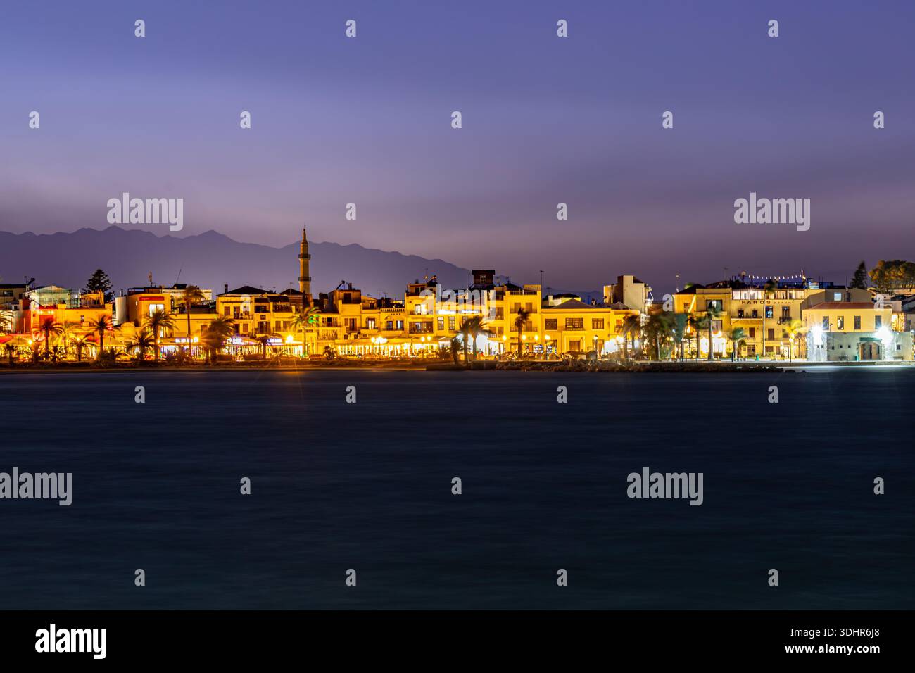 La vista di un affascinante paesaggio urbano si illumina al crepuscolo, mentre le luci brillano lungo il lungomare contrastando con il mare scuro, Rethimno, Rethymno, Gr Foto Stock