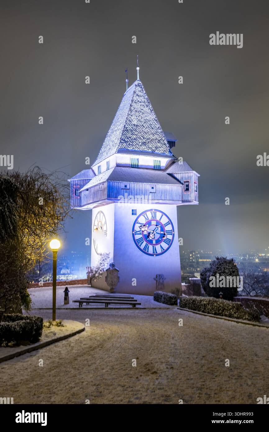 Vista della torre dell'orologio illuminata che si erge maestosamente contro il cielo notturno, i suoi dettagli spolverati di neve, che si illuminano dolcemente sopra la città, Graz, St Foto Stock