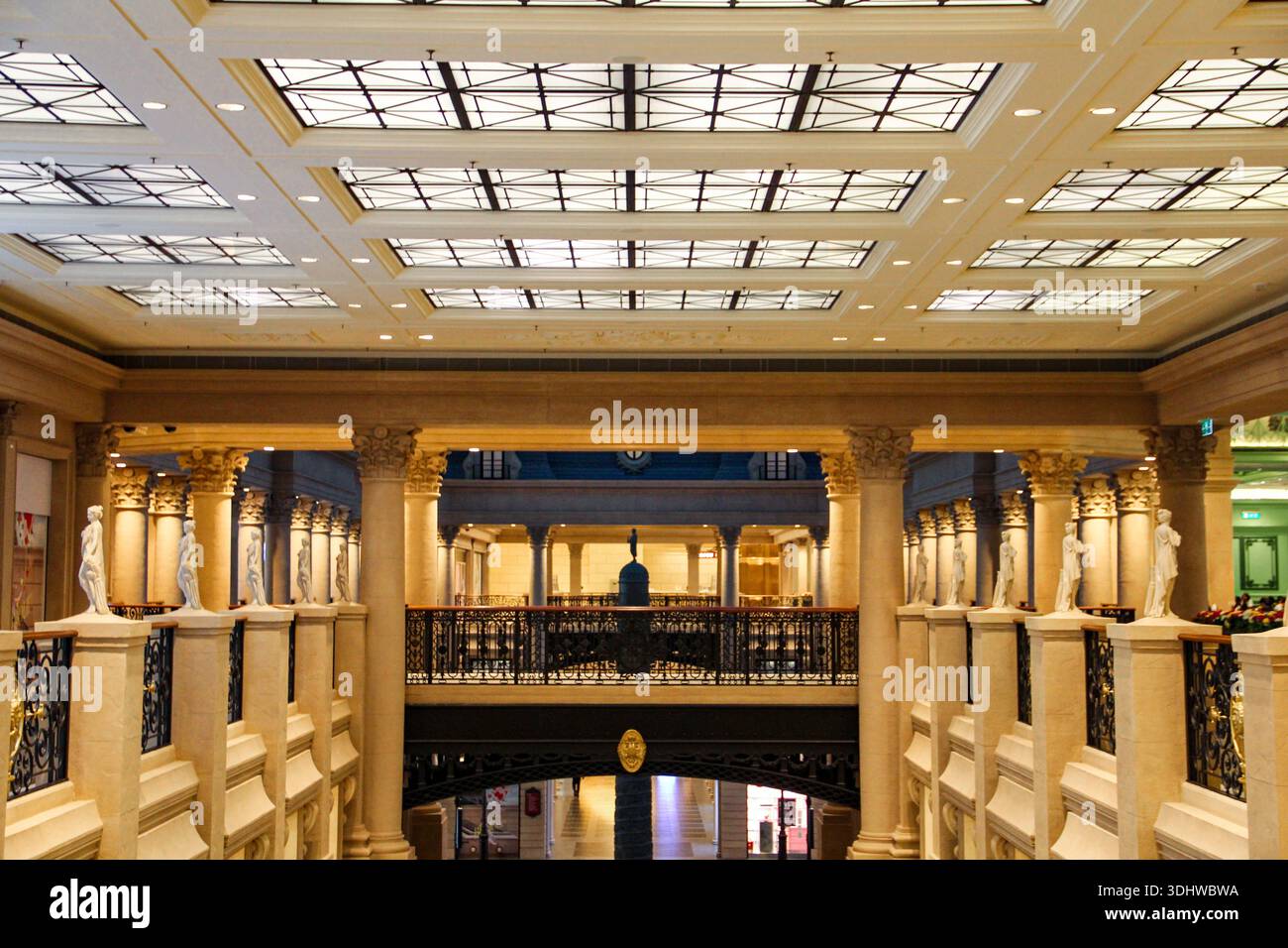 Vista interna simmetrica di una grande sala neoclassica con statue in marmo, colonne ornate e un soffitto geometrico lucernario in pietra beige. Foto Stock