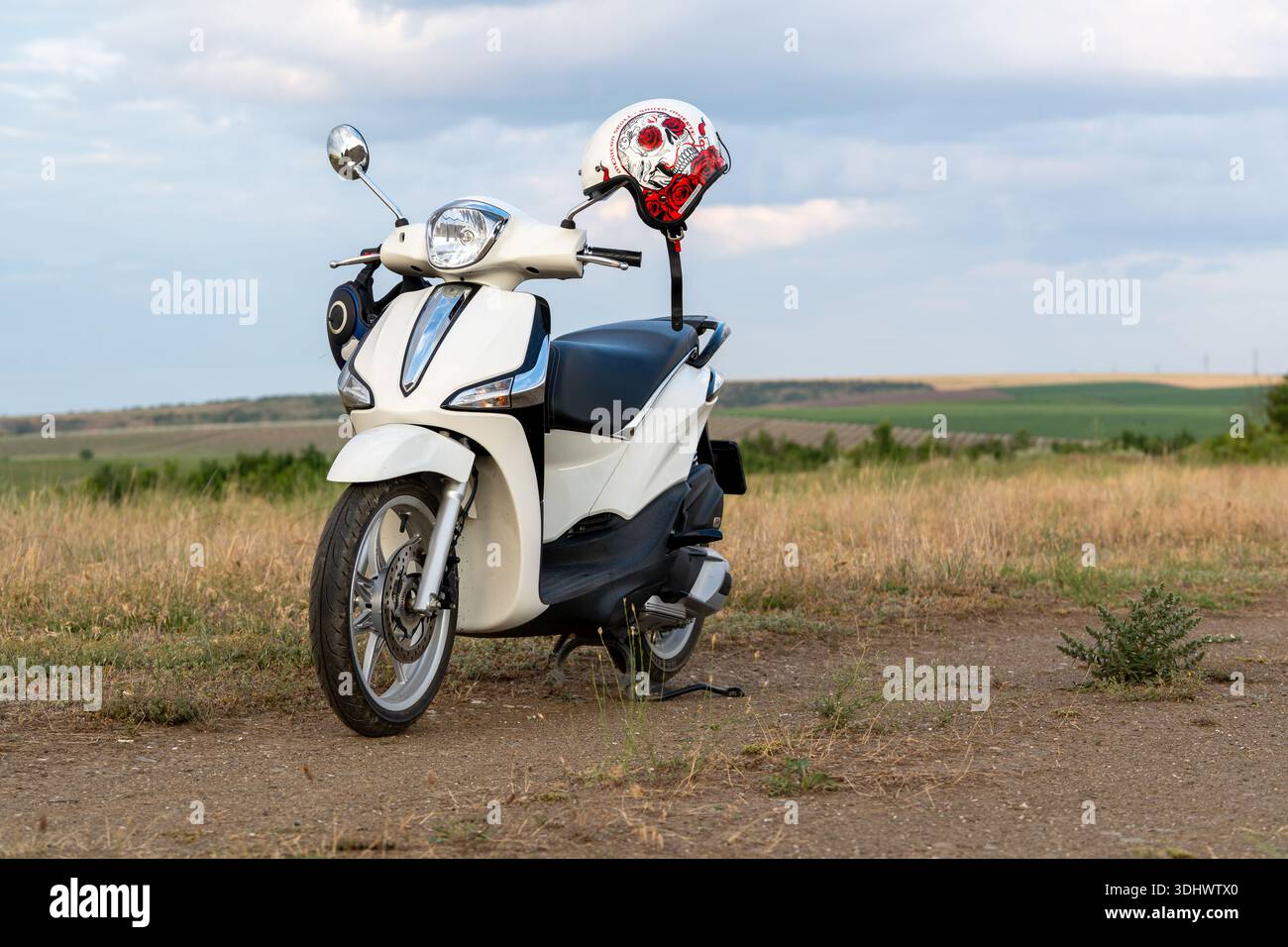 Uno scooter bianco si trova ancora su un sentiero sterrato in un campo erboso. Un casco poggia sul manubrio. Le verdi colline si estendono sullo sfondo sotto una S nuvolosa Foto Stock