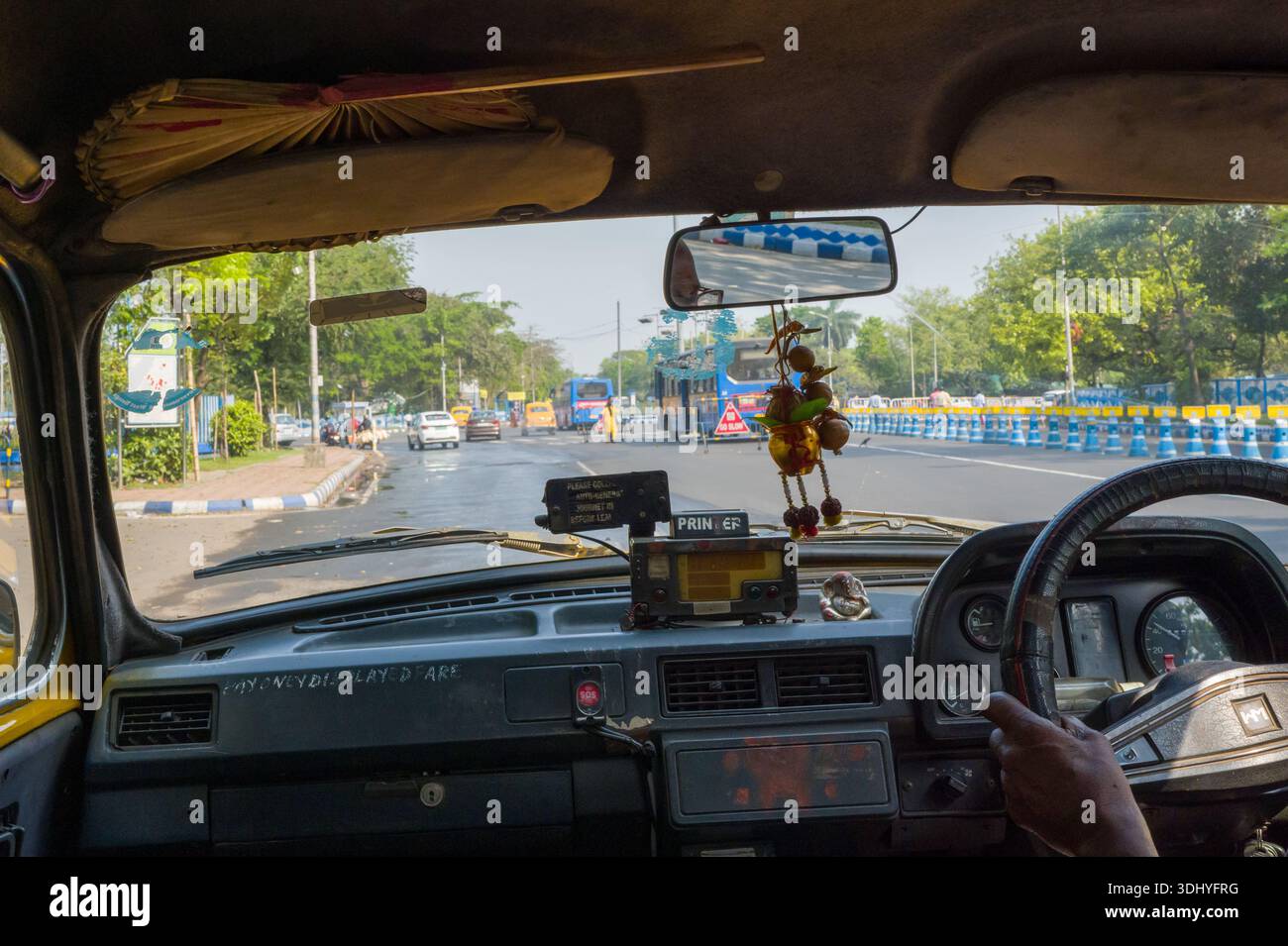 Calcutta, India - 24 marzo 2024: L'immagine mostra l'interno di un taxi Ambassador con la mano del conducente sul volante, i dettagli del cruscotto, il metro di tariffa, l'ornamento appeso e la vista attraverso il parabrezza di un'ampia strada urbana alberata con barriere blu e bianche ai bordi della strada a Calcutta. Foto Stock