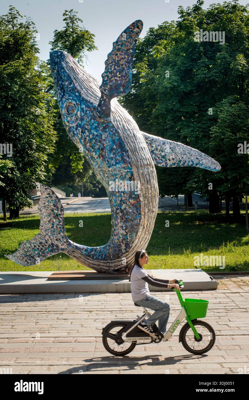 La Balena (la balena) scultura dell'artista italiano Jacopo Allegrucci, parte della 24a Mostra Internazionale della Triennale di Milano, commissionata nell'ambito della serie "la fragilità del futuro", l'enorme cartapesta blu m`chè cetacea è stata svelata il 13 maggio 2025, per simboleggiare la vulnerabilità delle specie in pericolo e la fragilità ecologica del mondo moderno. Fu incendiato il 14 luglio 2025 e ridotto a uno scheletro di metallo carbonizzato meno di un mese dopo la fotografia. Palazzo dell'Arte, Parco Sempione, Milano, Italia. Donna che passa in bicicletta a noleggio in città. Foto Stock