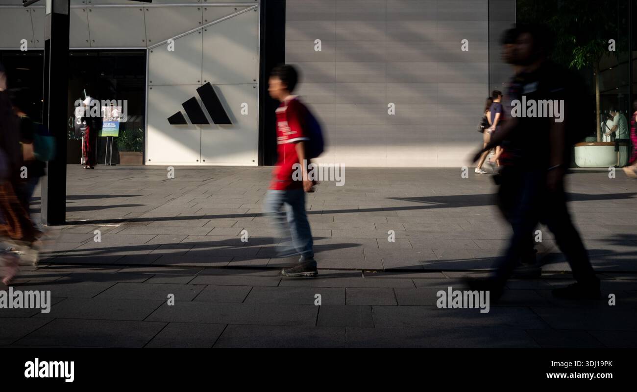 Suggestiva foto di strada davanti al negozio Adidas Singapore con logo e persone sfocate che camminano con un giovane in una camicia da calcio alla luce della sera Foto Stock