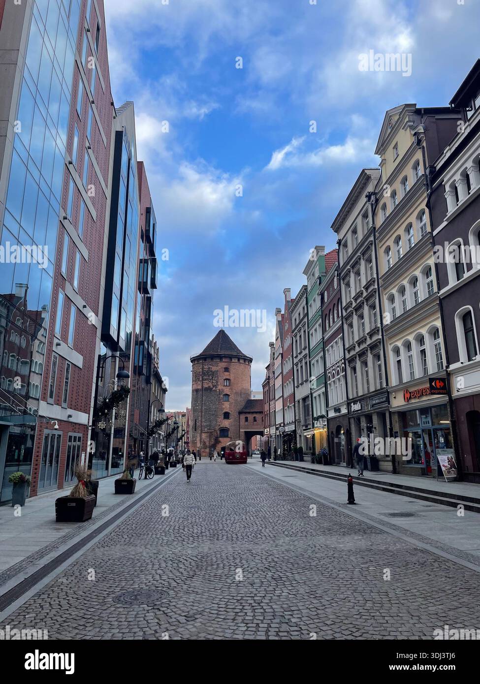 Strada acciottolata che conduce verso la storica porta della città nel centro storico di Gdańsk sotto un cielo blu, in parte nuvoloso. - Immagine stock catturata con smartphone