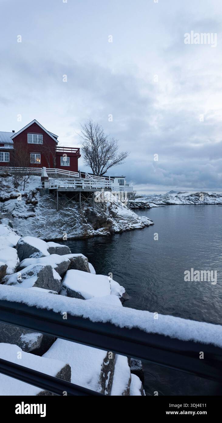 Svolver Norway Lofoten Islands Red House con un tetto bianco si trova su una collina che si affaccia su un corpo d'acqua. La casa è circondata da neve e rocce, e. Foto Stock