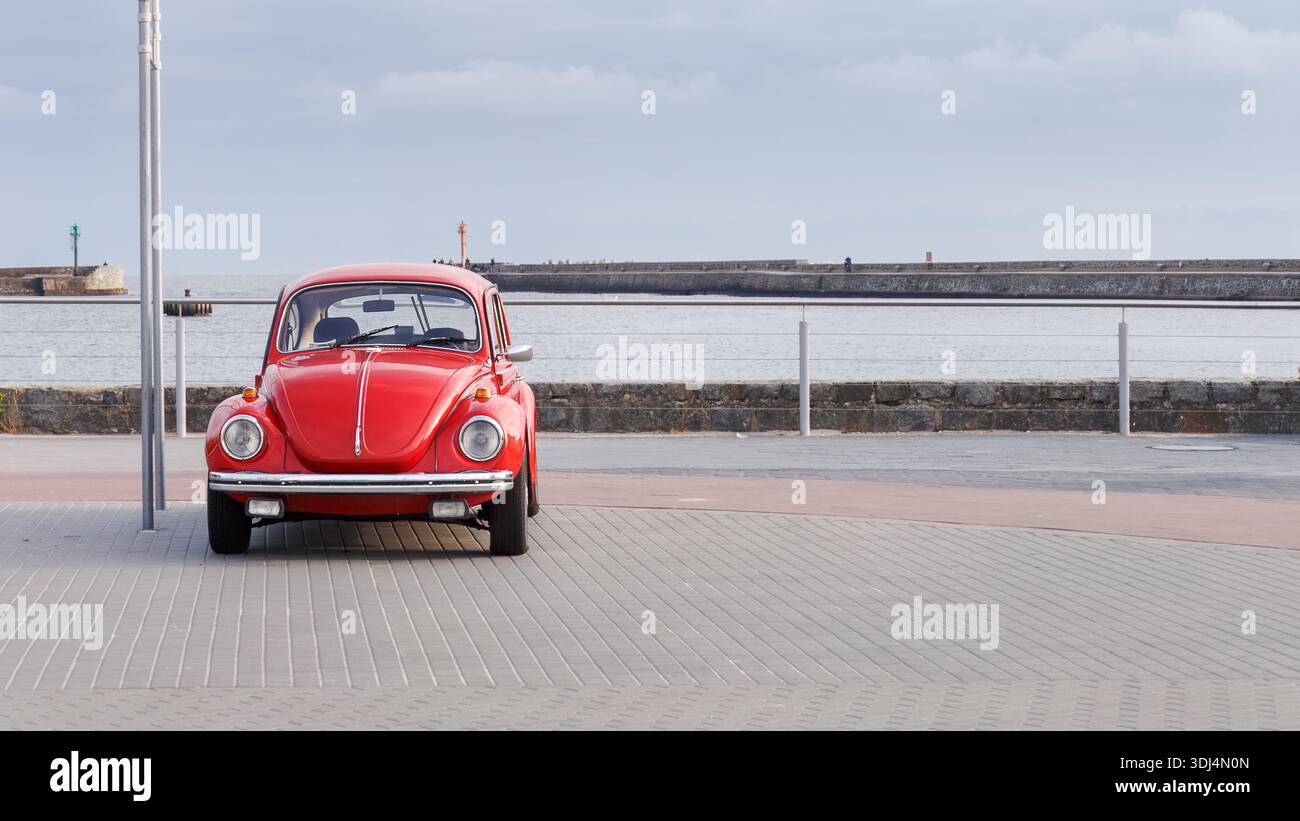 Fascino costiero con auto rossa d'epoca parcheggiata sulla passeggiata sul lungomare, orizzonte aperto del porto e luce soffusa d'estate che riflette la rilassata sommità lenta del Baltico Foto Stock