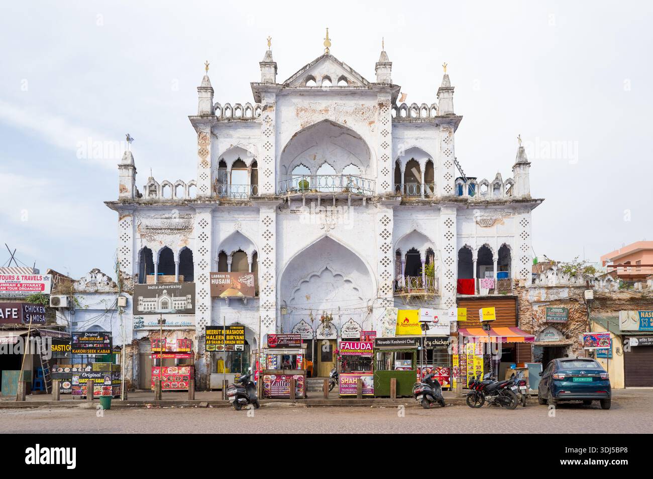Lucknow, India - 16 marzo 2024: L'immagine mostra uno storico edificio bianco con finestre ad arco ed elementi decorativi, con numerosi negozi a livello della strada con cartellonistica colorata, motociclette parcheggiate e un'auto davanti alla luce del giorno. Foto Stock