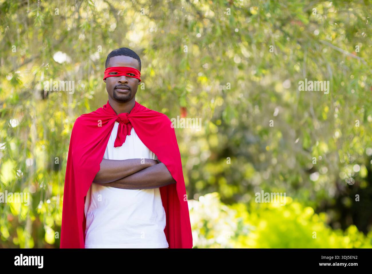 Uomo afro-americano in piedi al centro in un giardino soleggiato con maglietta bianca bendata rossa cape. Per esterni, ritratti, mantello, mascherato, fogliame, bokeh, sole Foto Stock