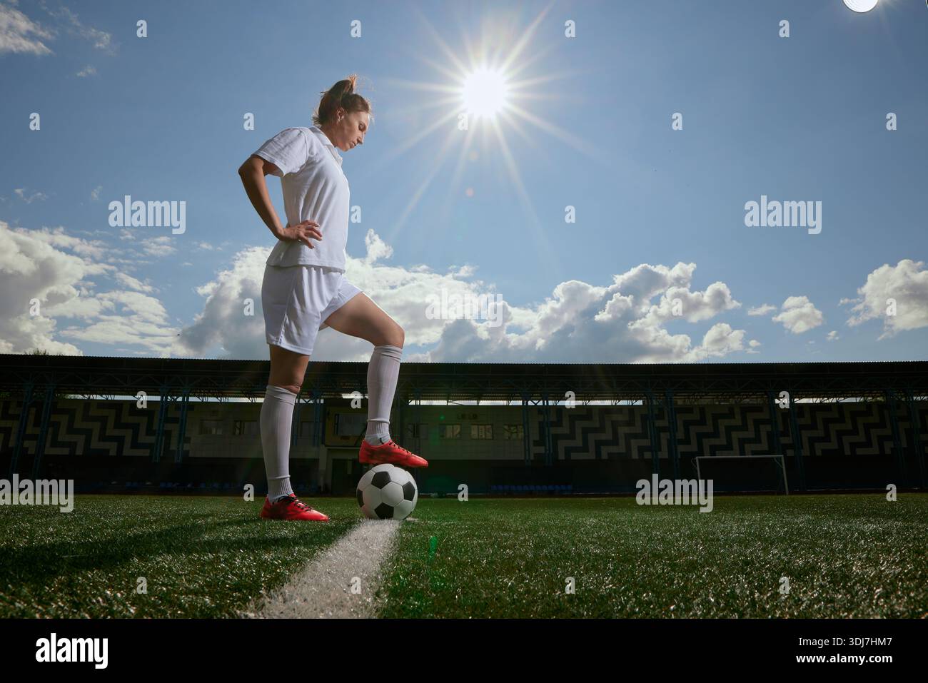 Sicura giocatrice di calcio con l'uniforme bianca, si erge con un piede sulla palla sotto il sole brillante sul campo. Foto Stock
