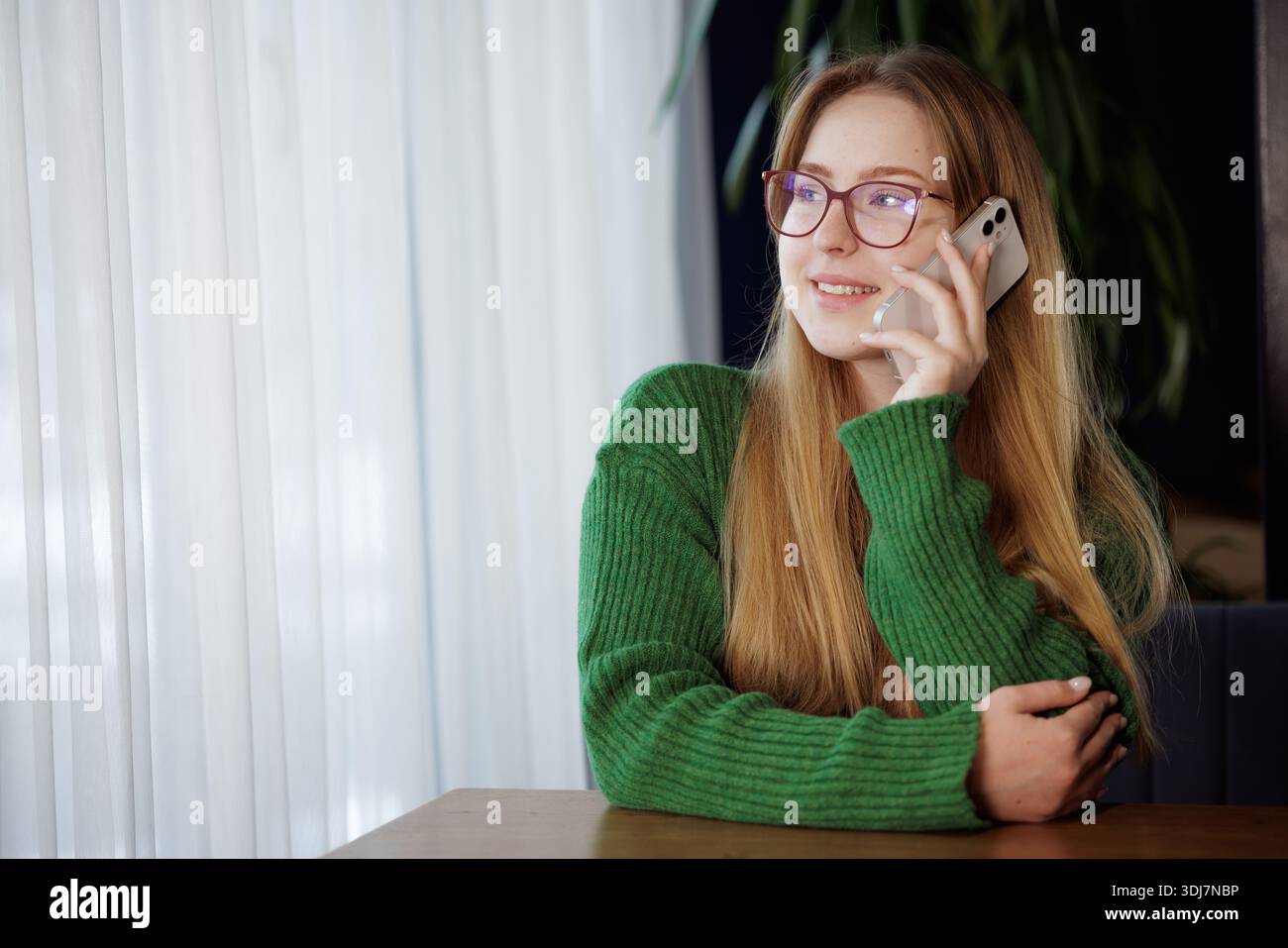 Sorridente giovane donna in occhiali che parla sullo smartphone mentre siede a un tavolo accanto alla finestra in un accogliente caffè. Foto Stock