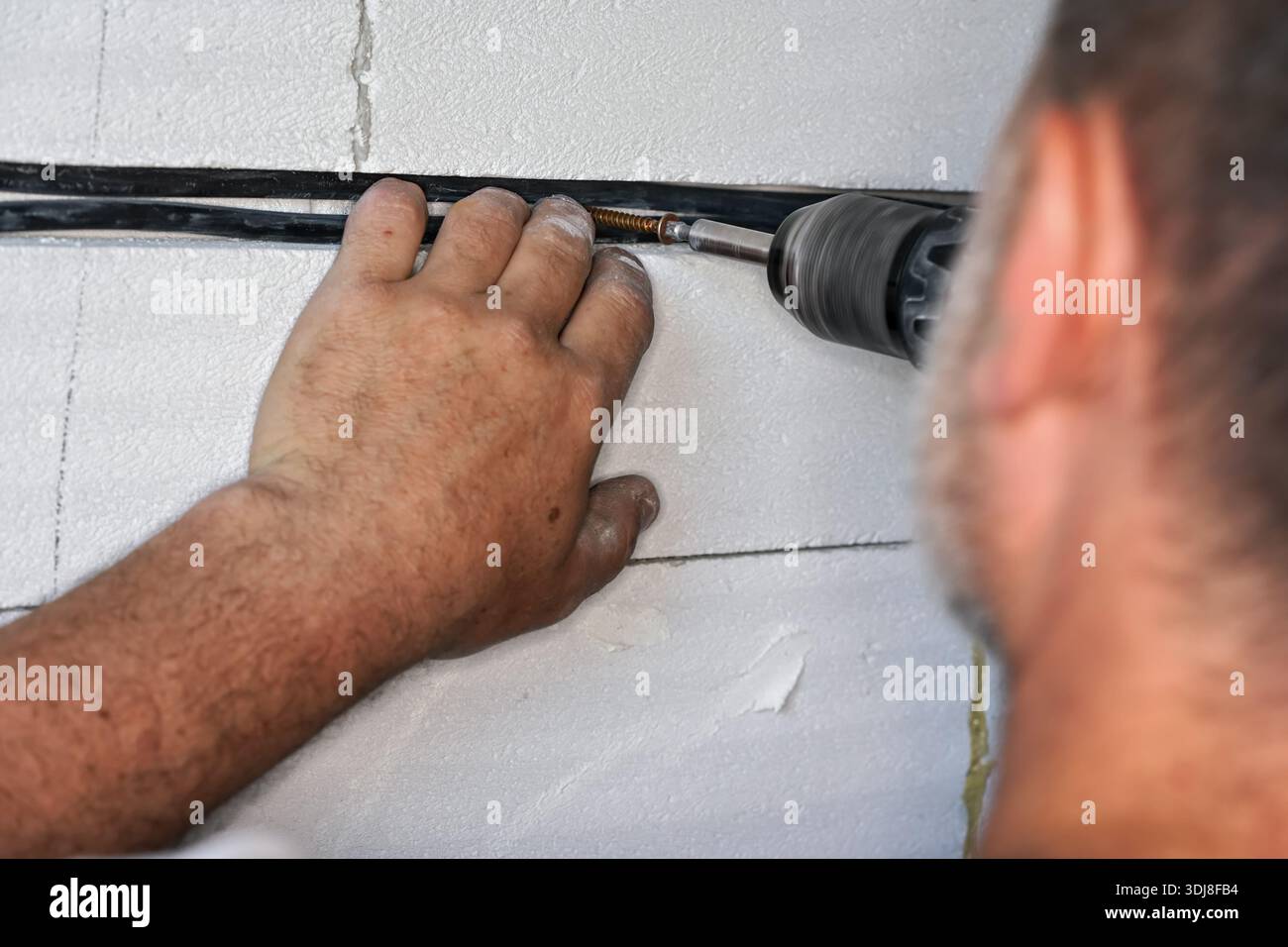 Elettricista che fissa i cavi nella scanalatura o inseguilo su una parete in una casa di nuova costruzione, utilizzando un trapano elettrico e viti - dettaglio dettagliato Foto Stock