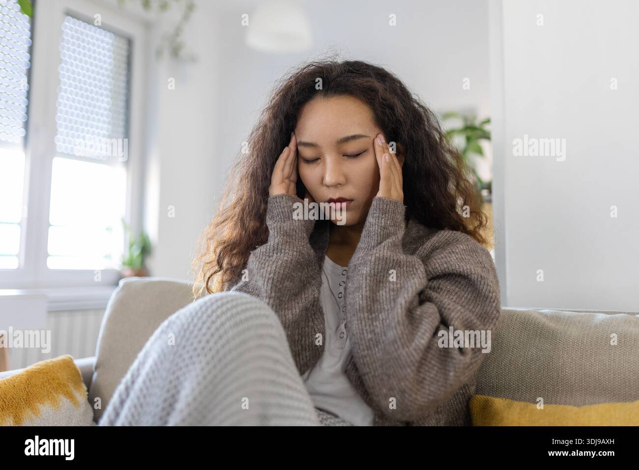 Ritratto di una giovane donna asiatica seduta sul divano a casa con mal di testa e dolore. Bella donna che soffre di mal di testa cronici quotidiani. Triste Wom Foto Stock