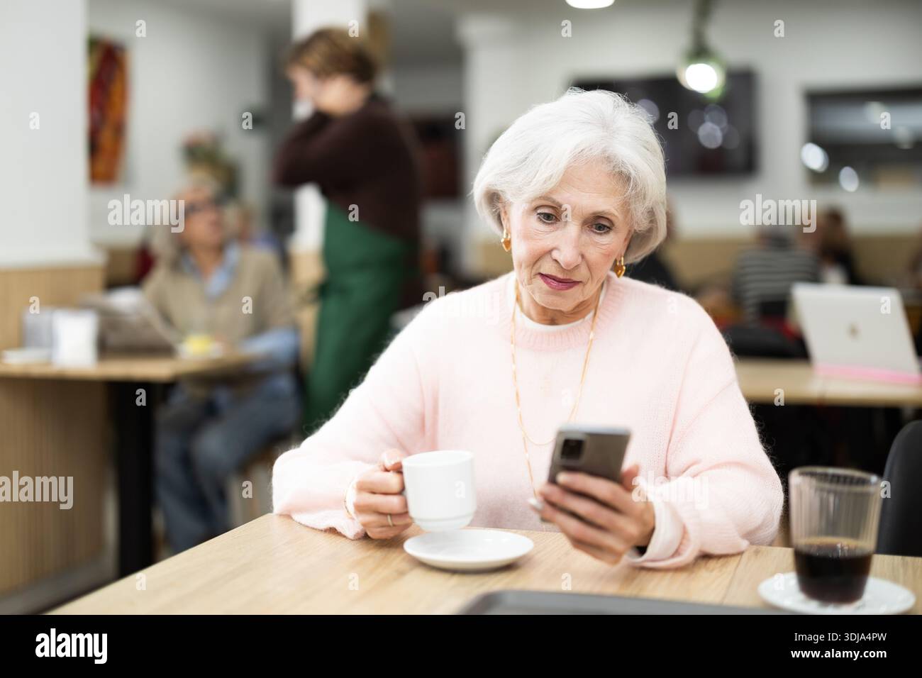 Una donna anziana in un bar beve caffè e usa il telefono cellulare Foto Stock